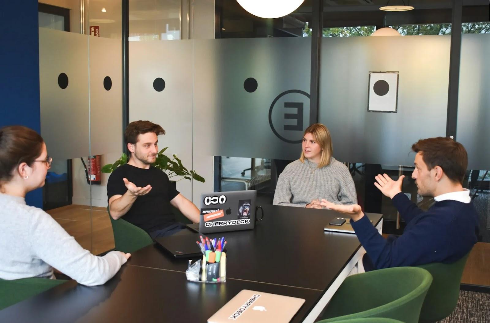 Four colleagues in green armchairs around a dark conference table, two men gesturing with open hands as they talk.
