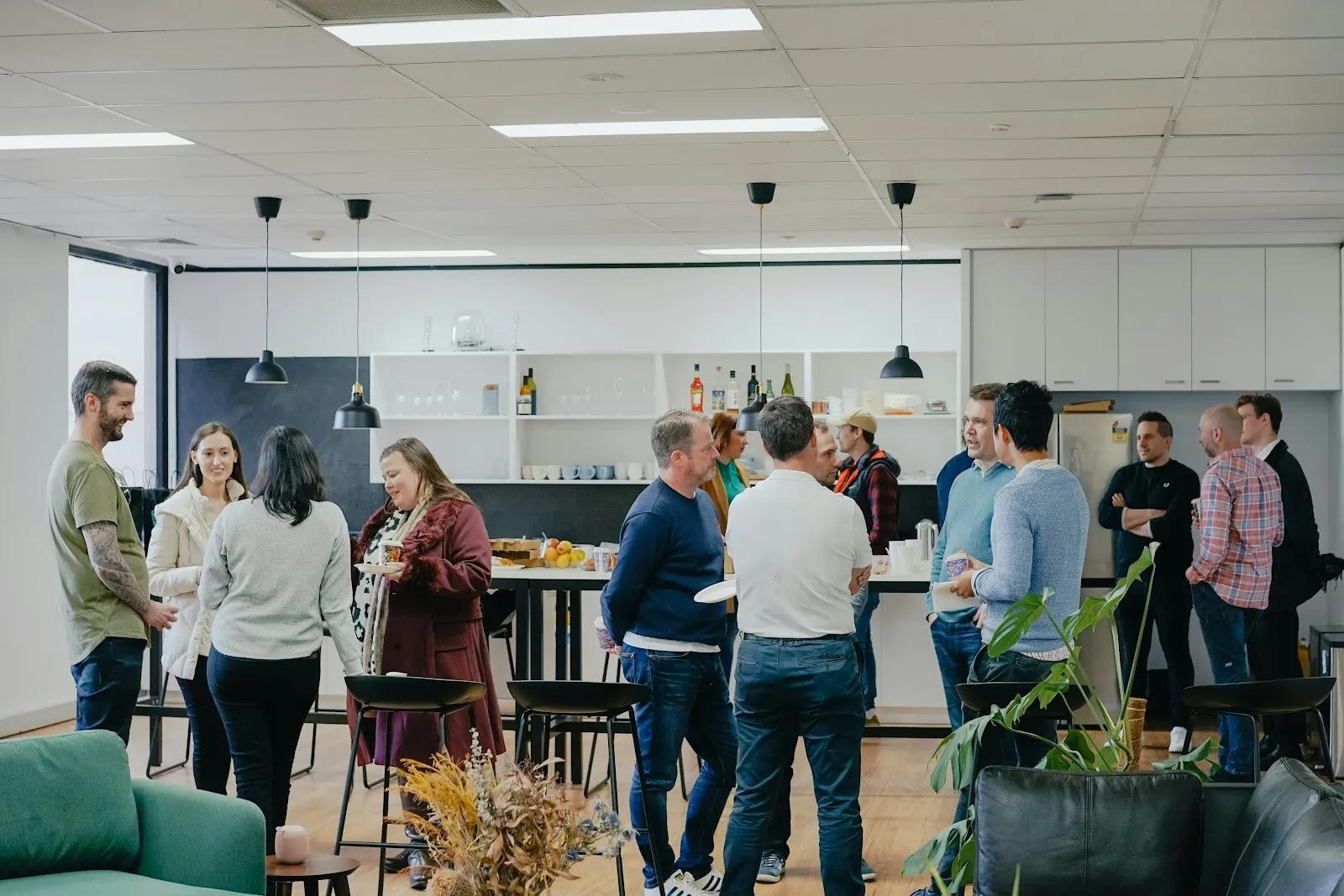 About a dozen people standing and talking in small groups in a bright office kitchen and lounge, one holding a paper plate.