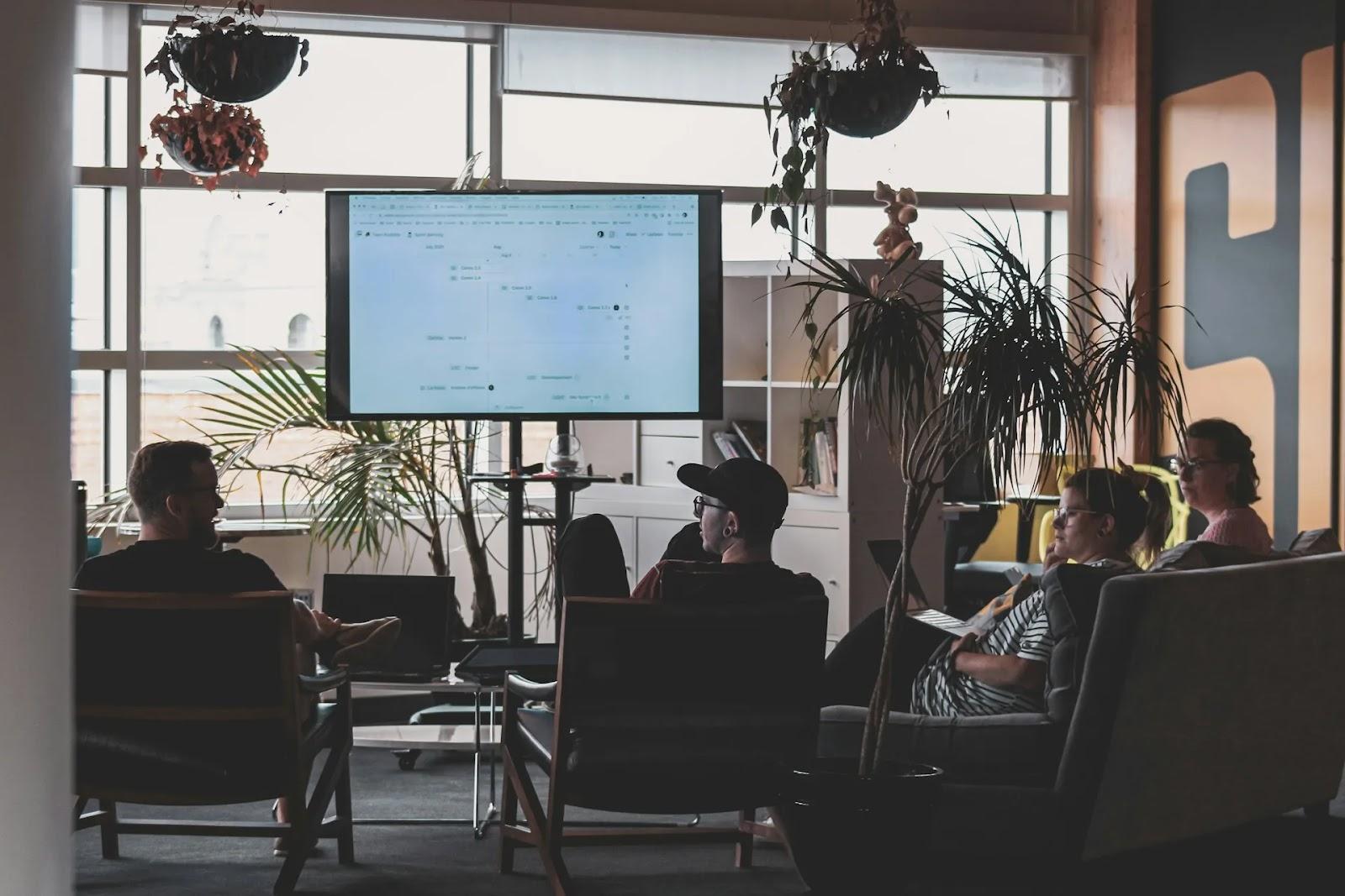Four people in armchairs and on a sofa watching a project timeline on a large screen in a bright office full of plants.