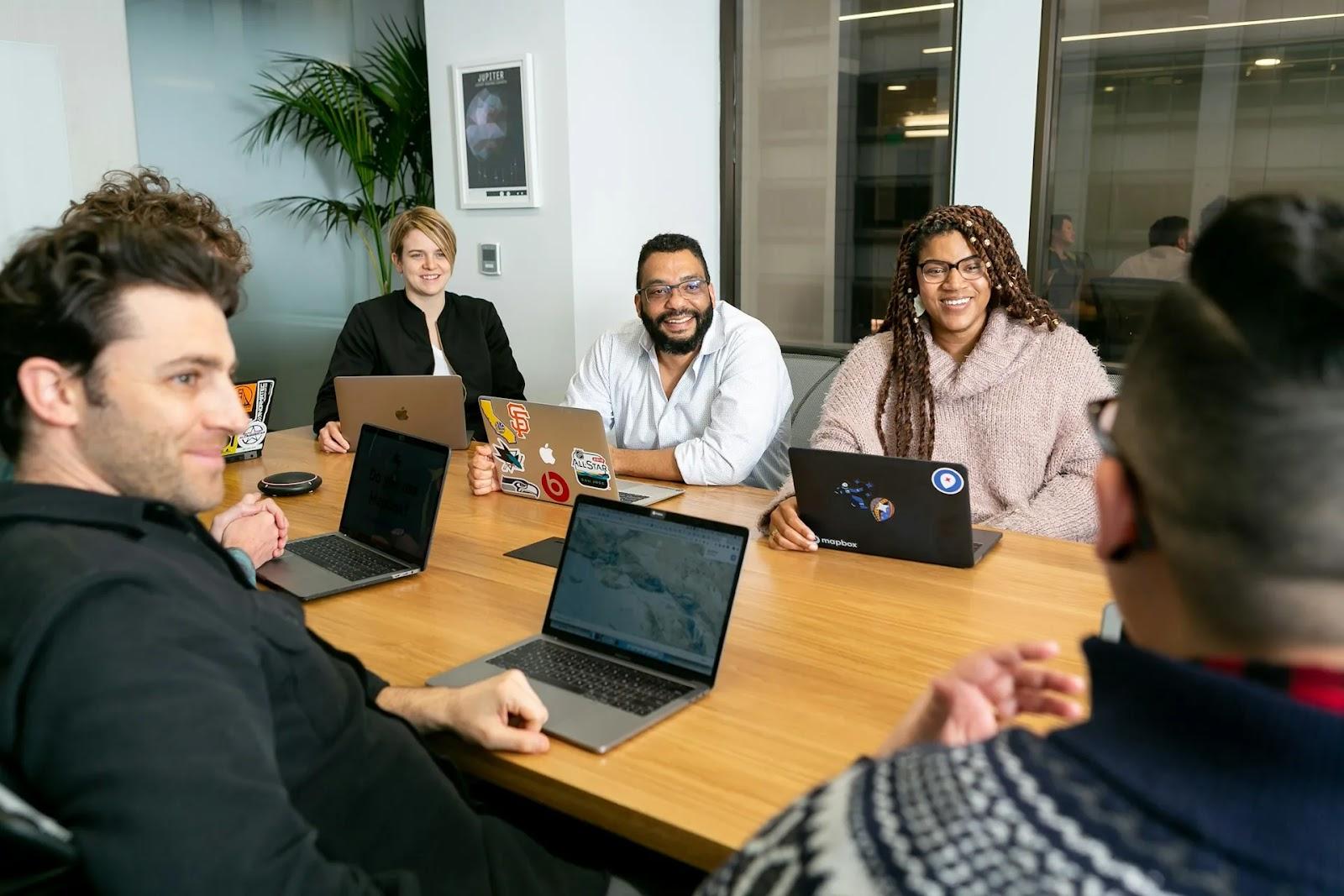 Six colleagues around a light wood conference table with open laptops, two of them smiling toward the camera.