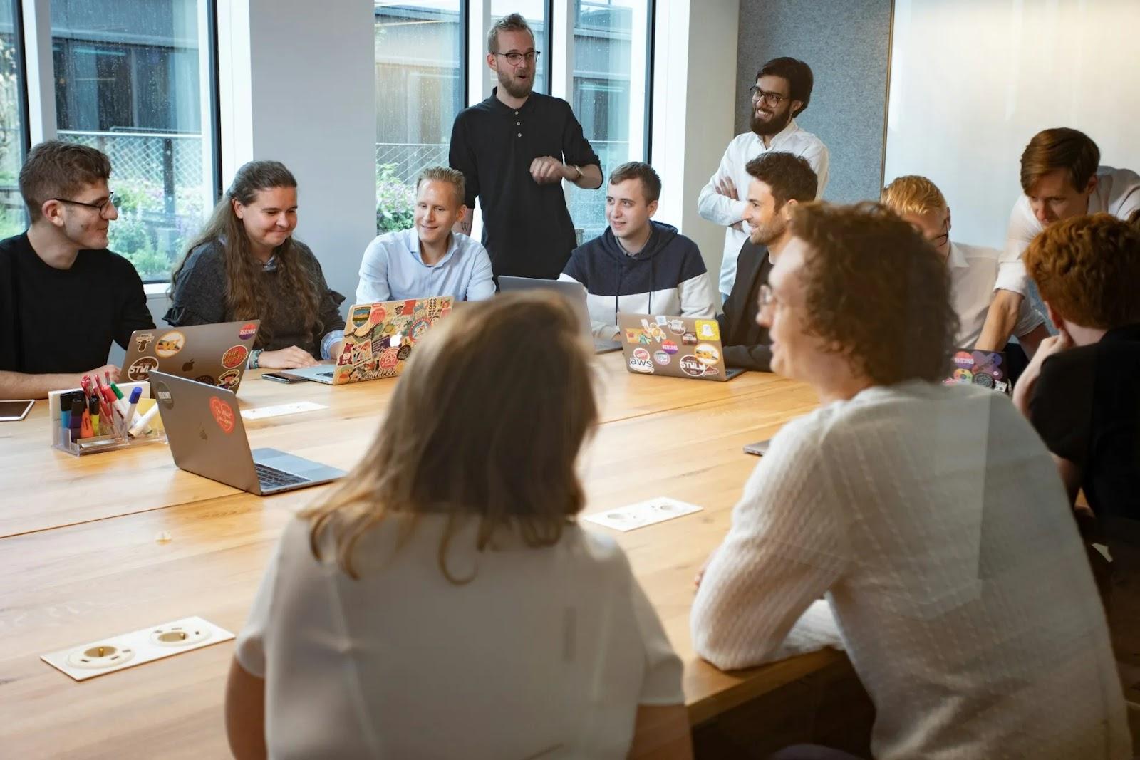 A bearded man standing by the windows and talking to about a dozen colleagues gathered around a long wooden table with laptops.