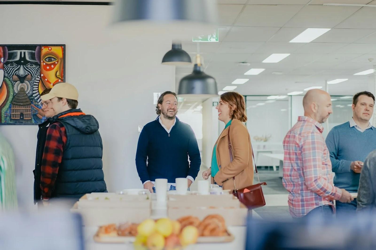 Small groups chatting in a bright office breakroom, with coffee and pastries on the counter and a colorful painting on the wall.