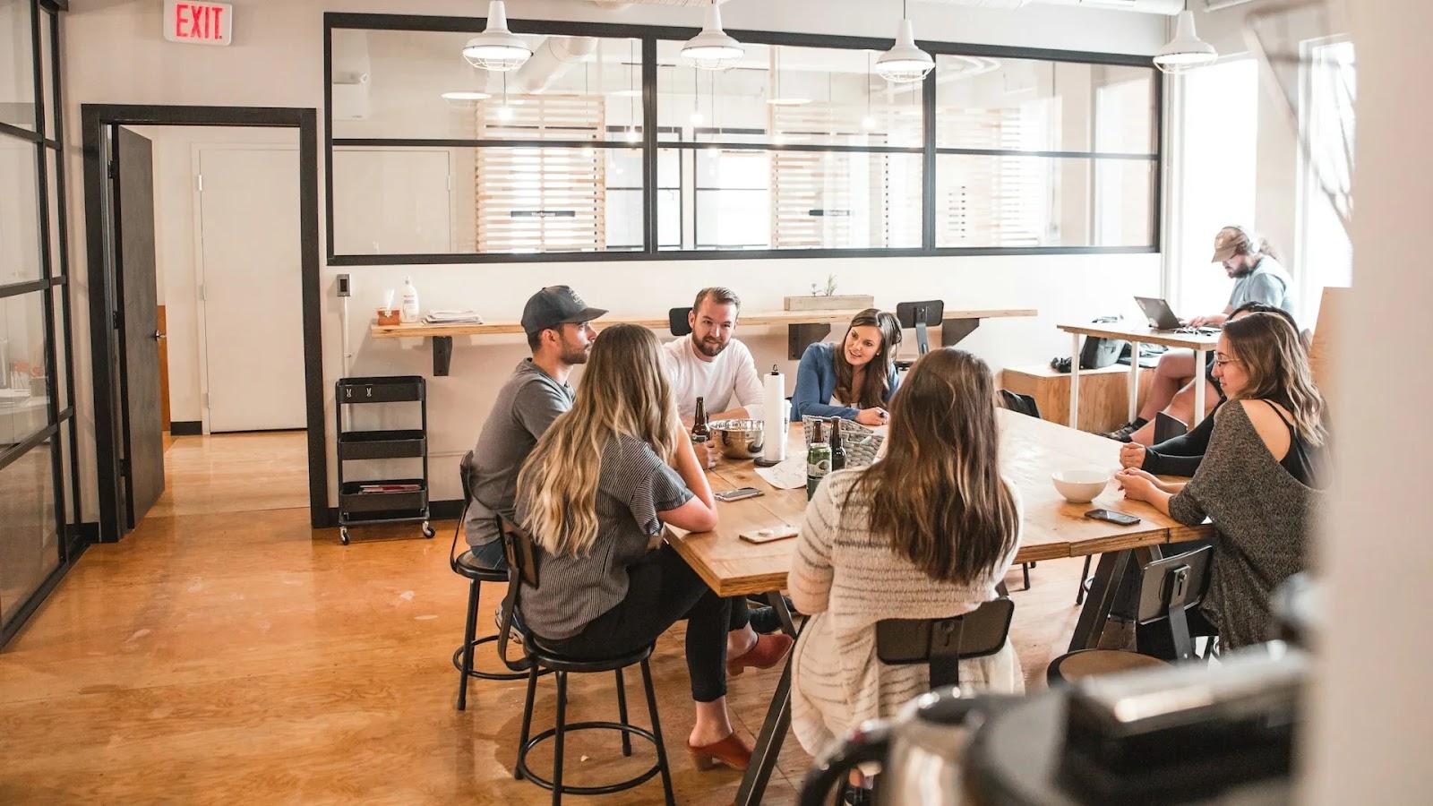 Seven people chatting around a long wooden communal table on black stools, with one person working at a desk by the window.