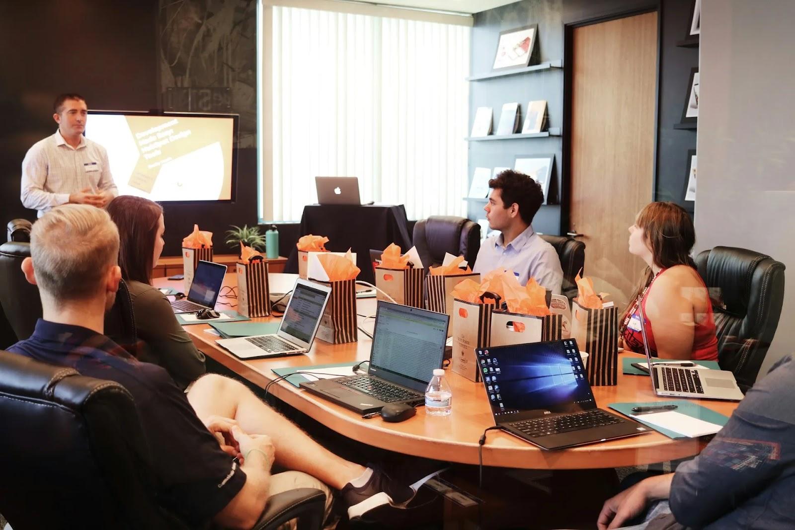 A man standing to present at the end of an oval conference table where seated guests have laptops and gift bags in front of them.