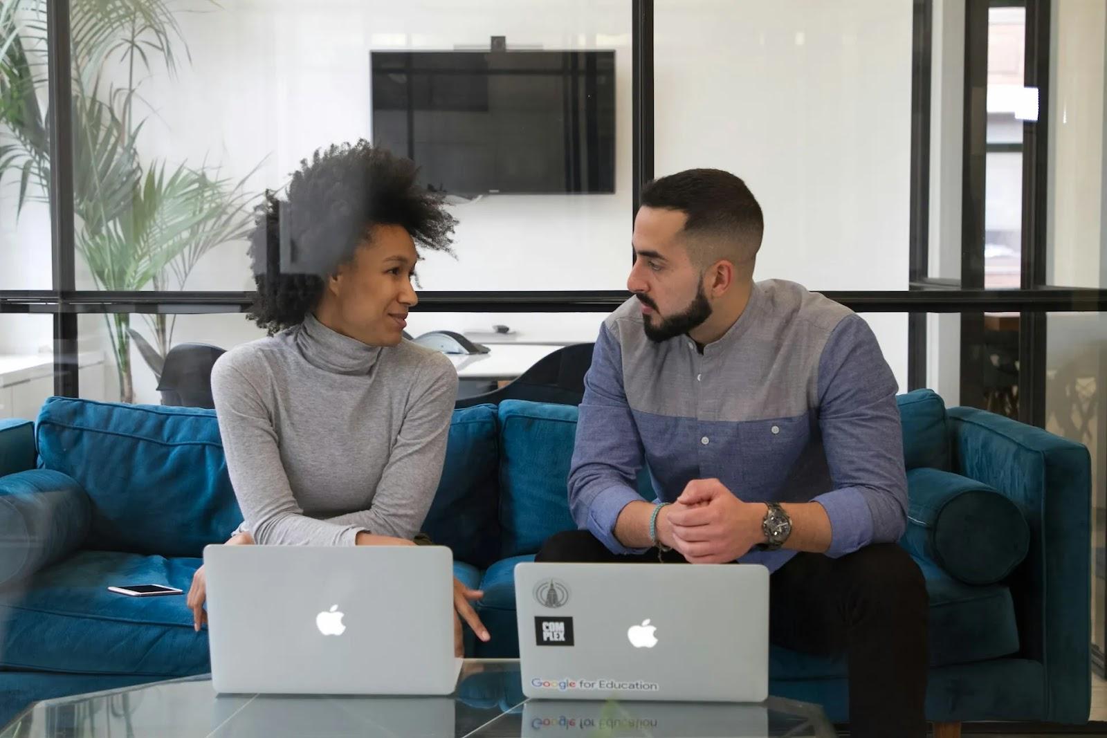 Two colleagues talking on a teal sofa behind a glass coffee table with two open laptops.