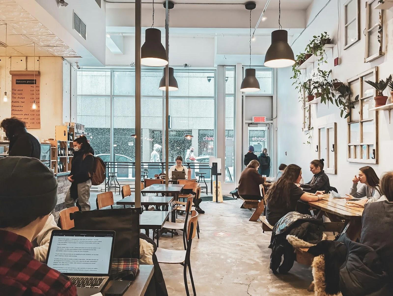 People working on laptops and talking at small wooden tables in a bright cafe with tall windows onto a snowy street.