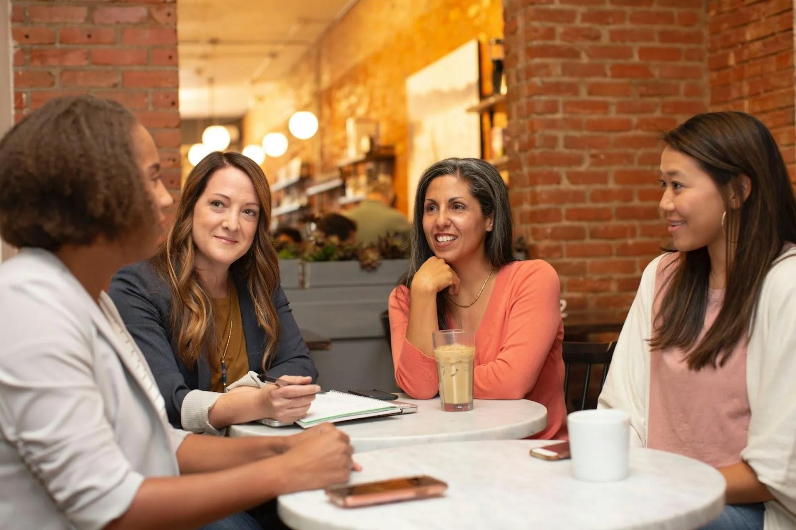 Four women talking around two small marble cafe tables by a brick wall, one taking notes in a notebook.
