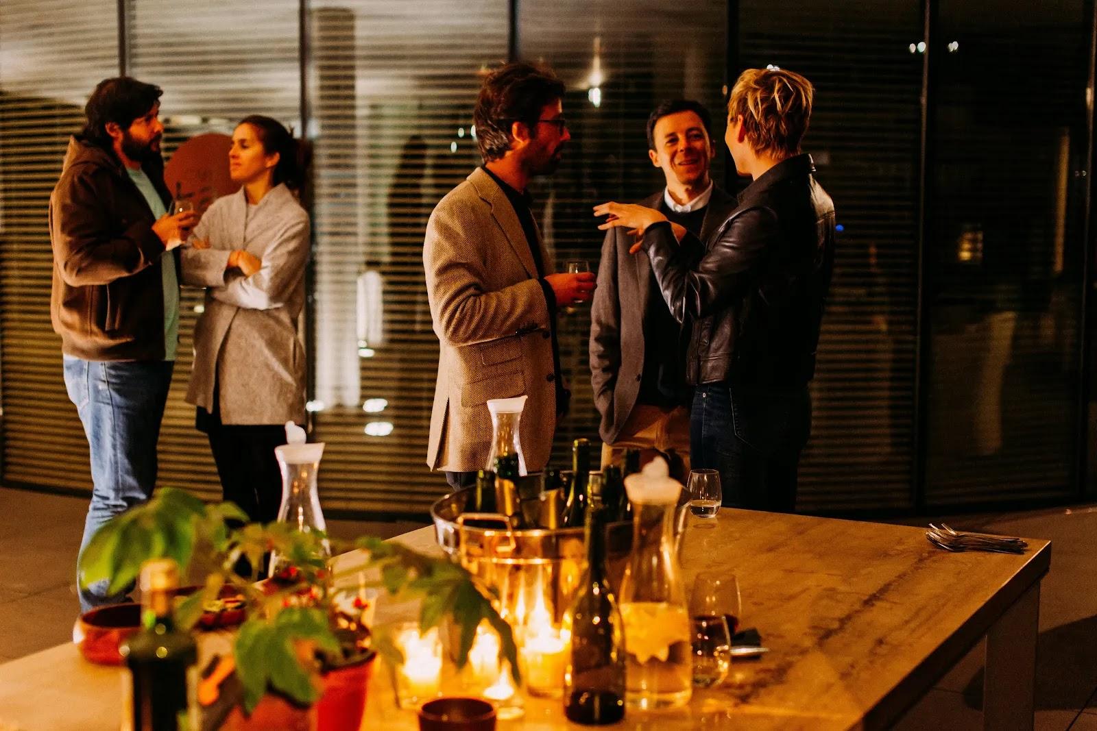 Five people standing and talking at an evening reception behind a candlelit table set with wine bottles and glasses.