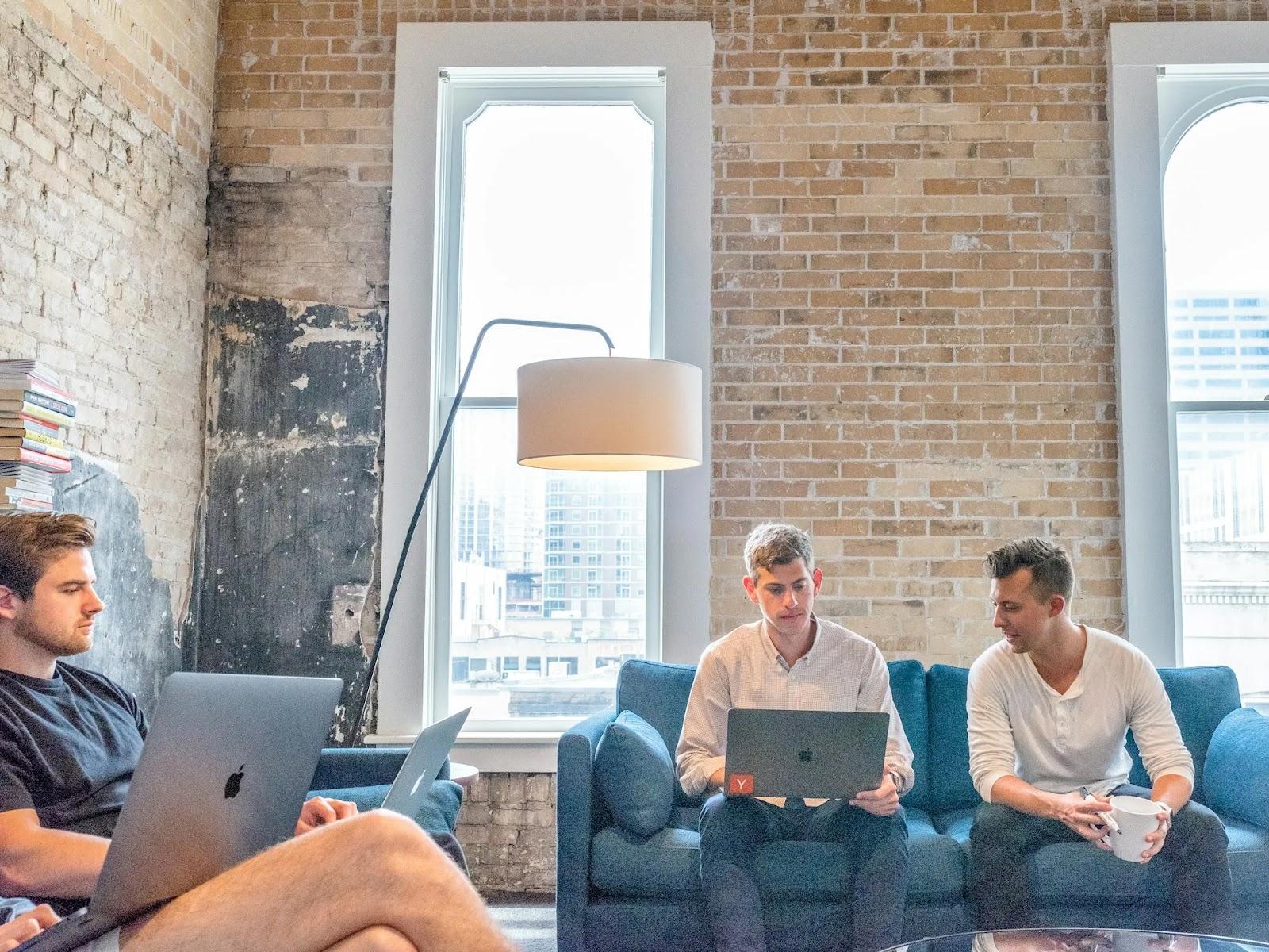 Three men on a teal sofa in a brick-walled loft, two working on laptops while the third holds a coffee mug and looks on.