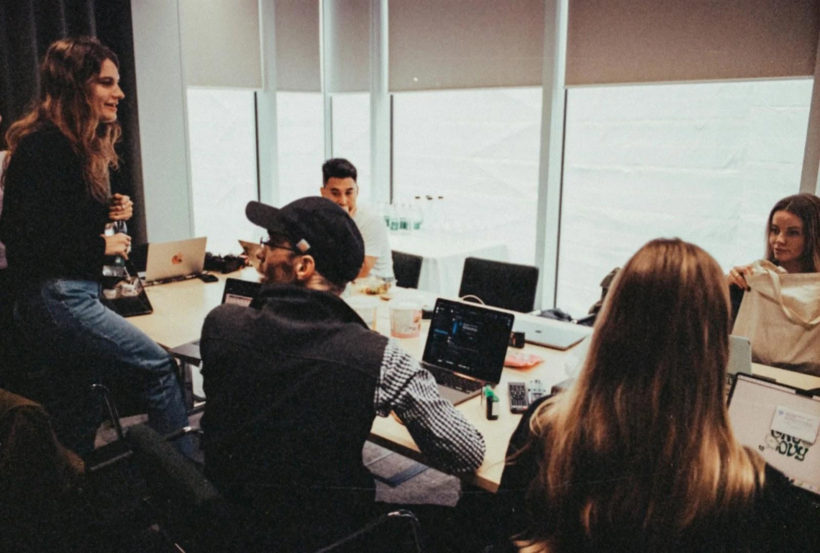 Five colleagues around a long conference table covered in laptops, one woman perched on the table edge as they talk.