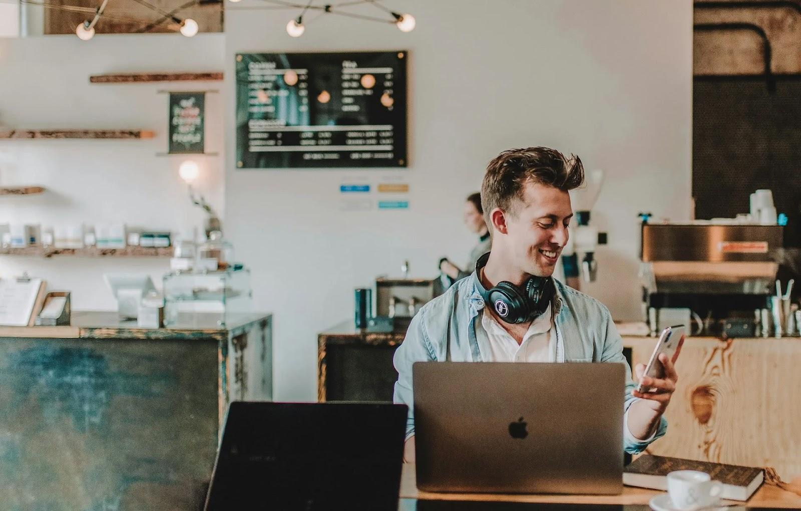 A young man smiling at his phone at a coffee shop table, with an open laptop and an espresso cup in front of him.