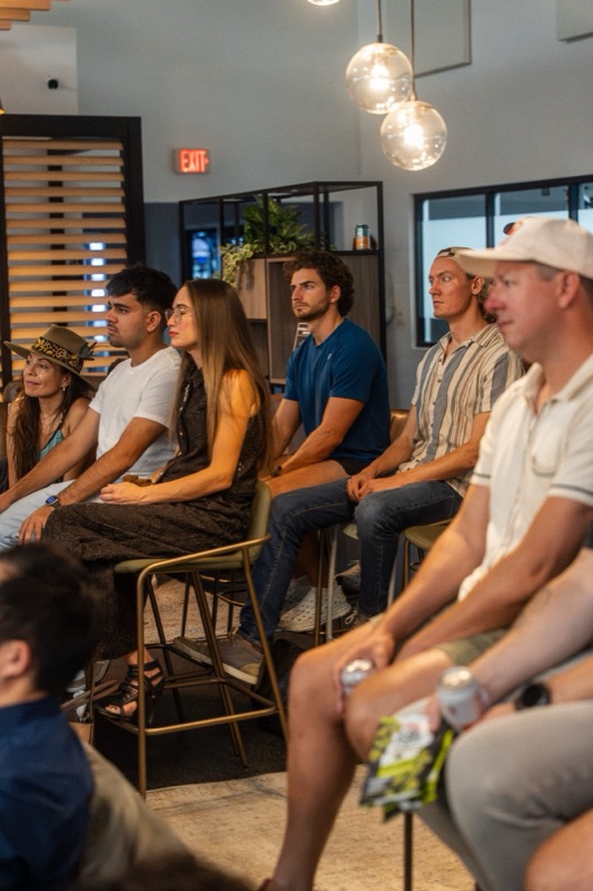 Attendees on bar stools watching a talk, seen from behind and the side, warm lounge light