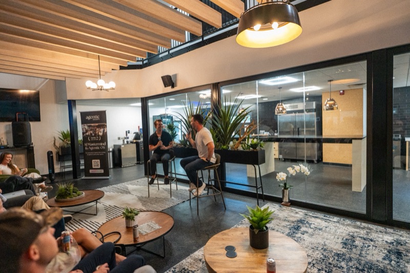 Two men in a fireside talk on stools, one gesturing, an AI Mastermind event banner behind them