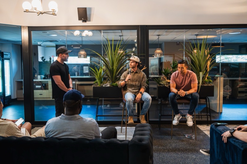 Two hosts on stools in a fireside talk with a standing moderator and a seated audience in a lounge