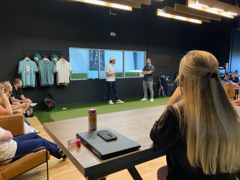 Two presenters standing in a golf-simulator studio talking to a seated group, retail shirts on the wall