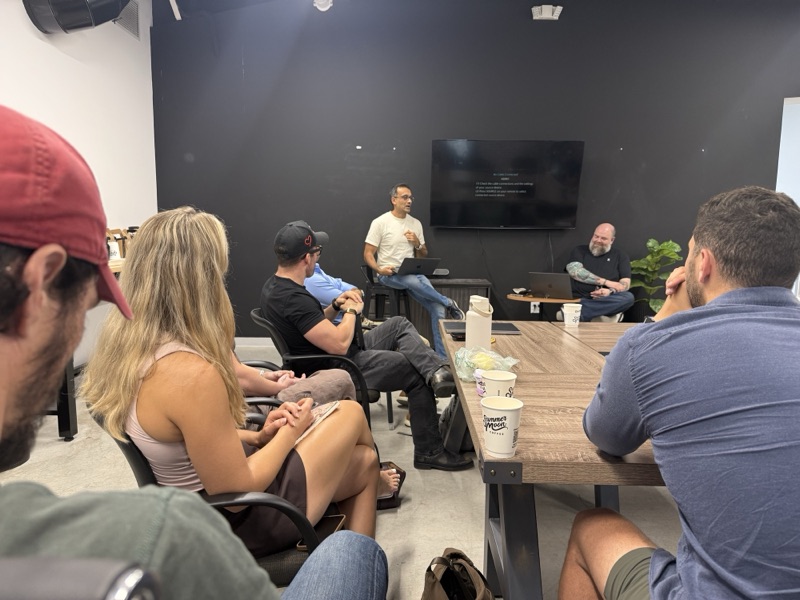Presenter seated on a stool addressing a small group in a dark-walled workshop room with a wall TV