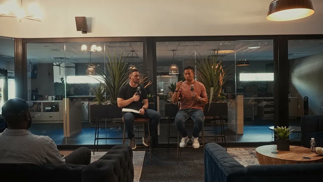 Connor Cullip and Mike Chu on stools in front of the glass wall at North Kollective in Austin, Mike holding a microphone