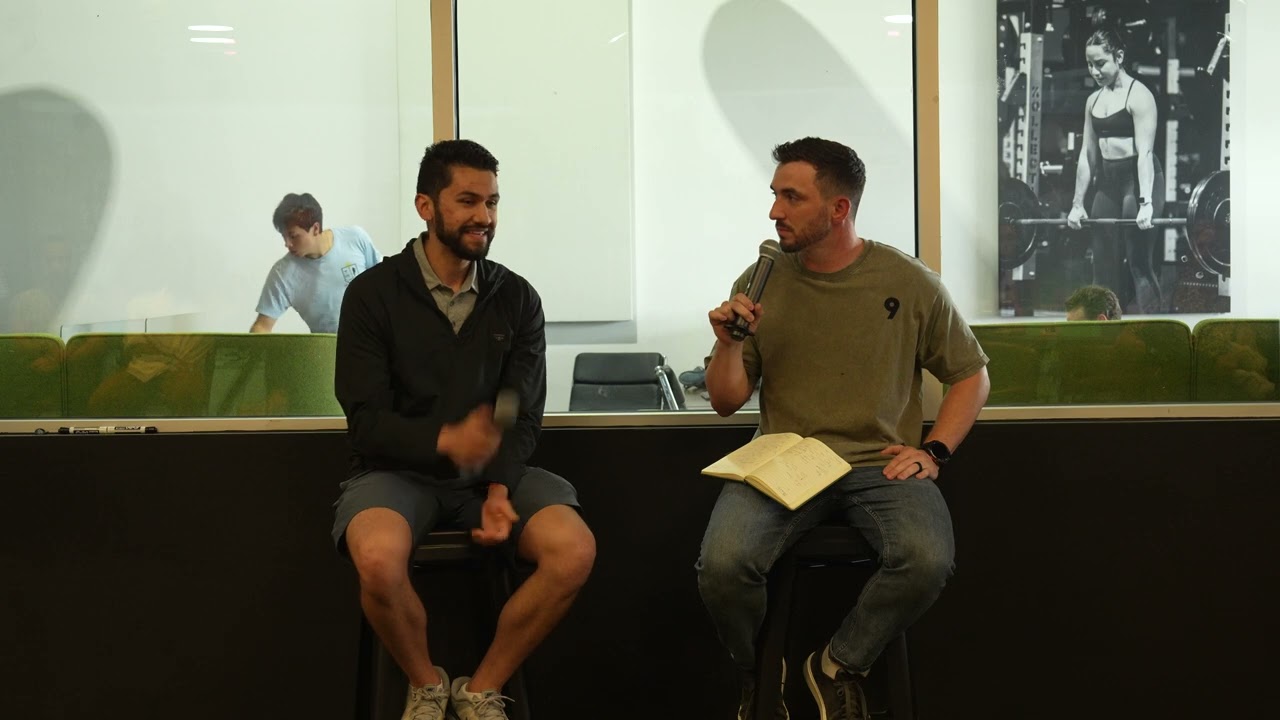 Alec Rios and Connor Cullip on tall stools in front of a glass wall, Connor holding a microphone and a notebook